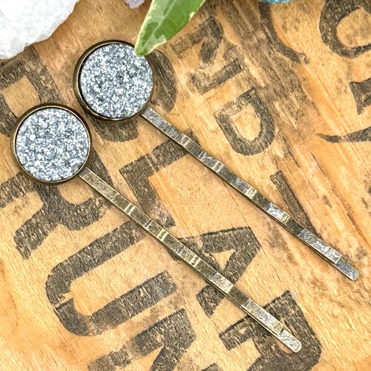 All Up In The Hair | Online Accessory Boutique Located in Mooresville, NC | Two Silver Glitter Druzy Bobby Pins on a wood background with black lettering. There are crystals and ivy leaves at the top of the image.