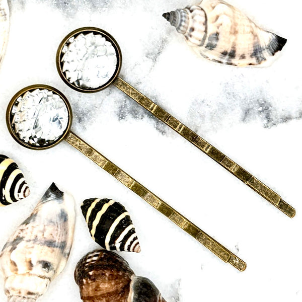 All Up In The Hair | Online Accessory Boutique Located in Mooresville, NC | Two Silver Mermaid Bobby Pins laying on a white marble background surrounded by shells.