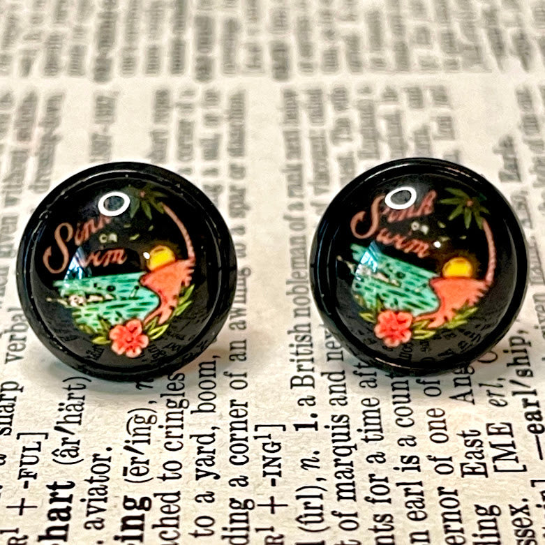 All Up In The Hair | Online Accessory Boutique Located in Mooresville, NC | Close up of two black earrings featuring a skeleton swimming in the ocean and the words "sink or swim". The earrings are laying on a book page.
