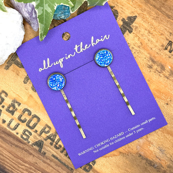 All Up In The Hair | Online Accessory Boutique Located in Mooresville, NC | Two Sky Blue Druzy Bobby Pins on an indigo colored, All Up In The Hair branded packaging card. The card is laying on a wood background with black lettering. There are crystals and ivy leaves at the top of the image. 