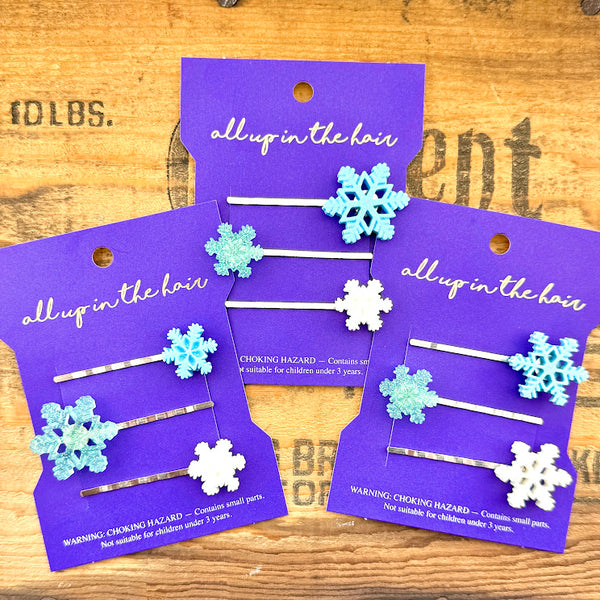 All Up In The Hair | Online Accessory Boutique Located in Mooresville, NC | Three sets of Snowflake Bobby Pins on an indigo colored, All Up In The Hair branded packaging card. The card is laying on a wood background with black lettering.