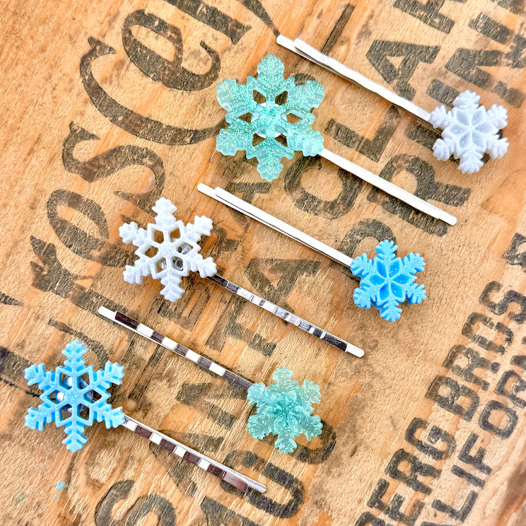 All Up In The Hair | Online Accessory Boutique Located in Mooresville, NC |All Up In The Hair | Online Accessory Boutique Located in Mooresville, NC | 6 Snowflake Bobby Pins on a wood background with black lettering. The snowflakes vary in color from light blue, mint, and white.