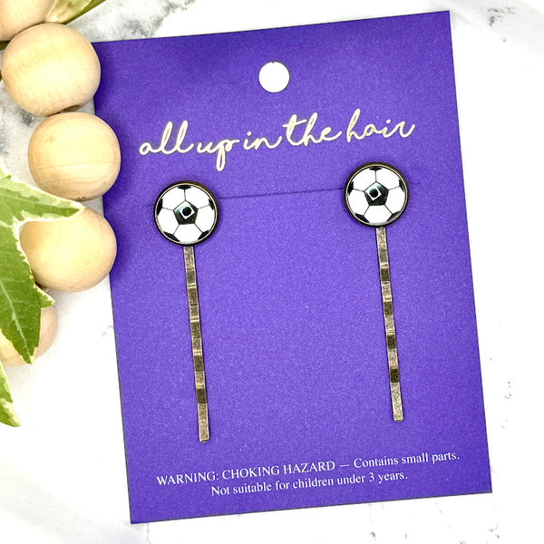 All Up In The Hair | Online Accessory Boutique Located in Mooresville, NC | Two Soccer Bobby Pins on an indigo colored, All Up In The Hair branded packaging card. The card is laying on a white marble background alongside a wood bead garland and ivy leaves.