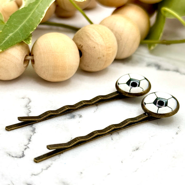 All Up In The Hair | Online Accessory Boutique Located in Mooresville, NC | Side view of two Soccer Bobby Pins laying on a white marble background alongside a wood bead garland and ivy leaves.