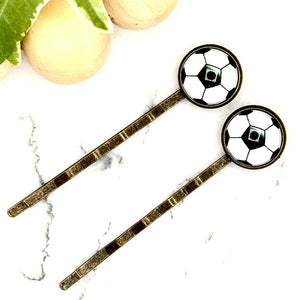 All Up In The Hair | Online Accessory Boutique Located in Mooresville, NC | Two Soccer Bobby Pins laying on a white marble background alongside a wood bead garland and ivy leaves.