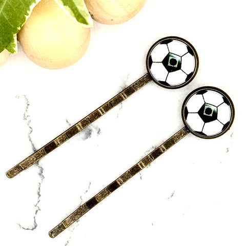 All Up In The Hair | Online Accessory Boutique Located in Mooresville, NC | Two Soccer Bobby Pins laying on a white marble background alongside a wood bead garland and ivy leaves.