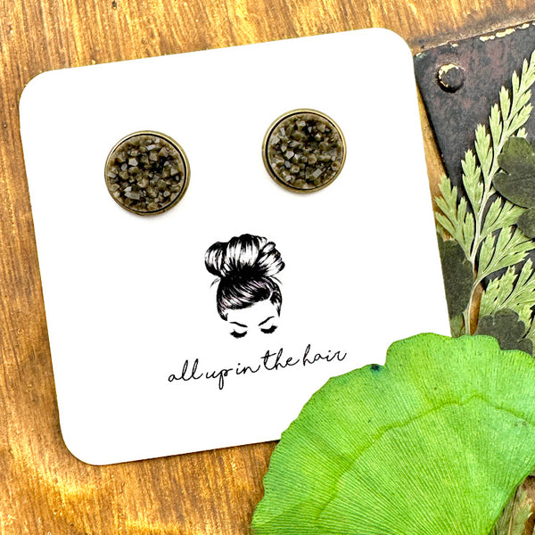 All Up In The Hair | Online Accessory Boutique Located in Mooresville, NC | Two dark grey druzies set in bronze settings. The earrings are on a white, All Up In The Hair branded packaging card. The card is laying on a wood background alongside green leaves.