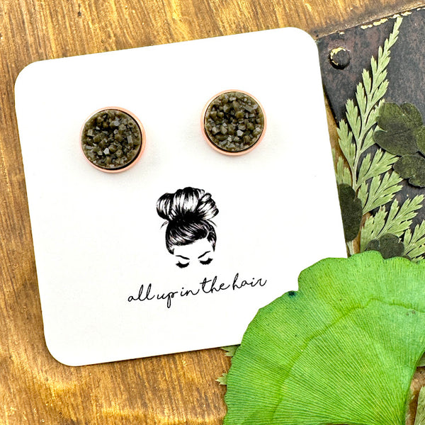 All Up In The Hair | Online Accessory Boutique Located in Mooresville, NC | Two dark grey druzies set in rose gold settings. The earrings are on a white, All Up In The Hair branded packaging card. The card is laying on a wood background alongside green leaves.