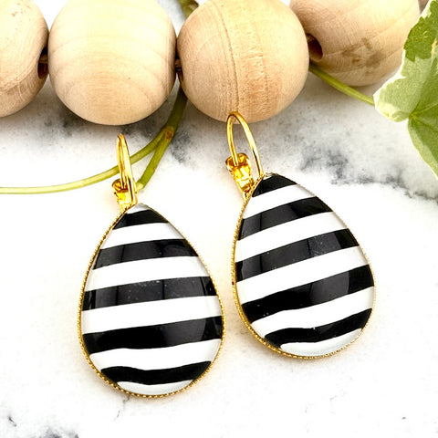 All Up In The Hair | Online Accessory Boutique Located in Mooresville, NC | Two black and white striped teardrop earrings on a white marble background. Behind the earrings is a wood bead garland and ivy leaves.
