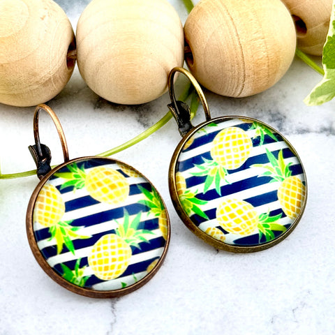 All Up In The Hair | Online Accessory Boutique Located in Mooresville, NC | Two round earrings with navy blue stripes and yellow pineapples on a white marble background. Behind the earrings is a wood bead garland and ivy leaves.