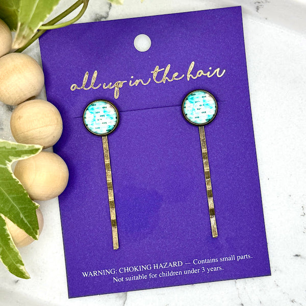 All Up In The Hair | Online Accessory Boutique Located in Mooresville, NC | Two Teal Arrow Bobby Pins on an indigo colored, All Up In The Hair branded packaging card. The card is laying on a white marble background alongside a wood bead garland and ivy leaves.
