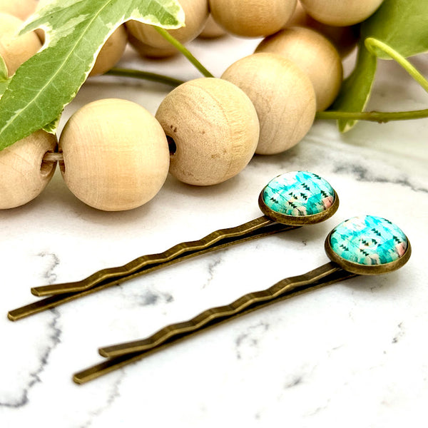 All Up In The Hair | Online Accessory Boutique Located in Mooresville, NC | Side view of two Teal Arrow Bobby Pins on a white marble background alongside a wood bead garland and ivy leaves.