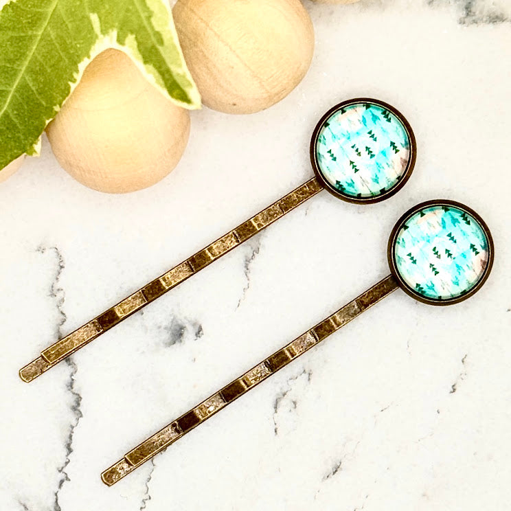 All Up In The Hair | Online Accessory Boutique Located in Mooresville, NC | Two Teal Arrow Bobby Pins laying diagonally on a white marble background alongside a wood bead garland and ivy leaves.