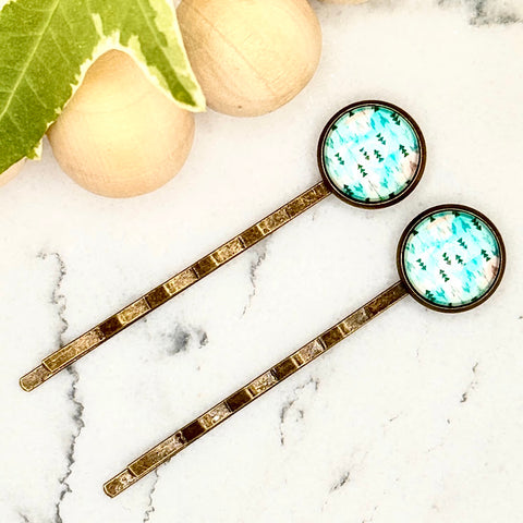 All Up In The Hair | Online Accessory Boutique Located in Mooresville, NC | Two Teal Arrow Bobby Pins laying diagonally on a white marble background alongside a wood bead garland and ivy leaves.
