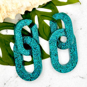 All Up In The Hair | Online Accessory Boutique Located in Mooresville, NC | Two Teal Chain Earrings laying on two monstera leaves on a white marble background.