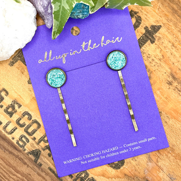 All Up In The Hair | Online Accessory Boutique Located in Mooresville, NC | Two Teal Druzy Bobby Pins on an indigo colored, All Up In The Hair branded packaging card. The card is laying on a wood background with black lettering. There are crystals and ivy leaves at the top of the image.