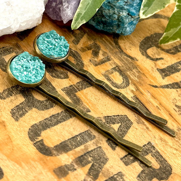 All Up In The Hair | Online Accessory Boutique Located in Mooresville, NC | Side view of our teal druzy bobby pins on a wood background with black lettering. There are crystals and ivy leaves at the top of the image.