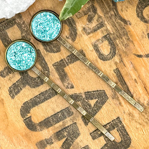 All Up In The Hair | Online Accessory Boutique Located in Mooresville, NC | Two Teal Druzy Bobby Pins laying diagonally on a wood background with black lettering. There are crystals an ivy leaves at the top of the image.