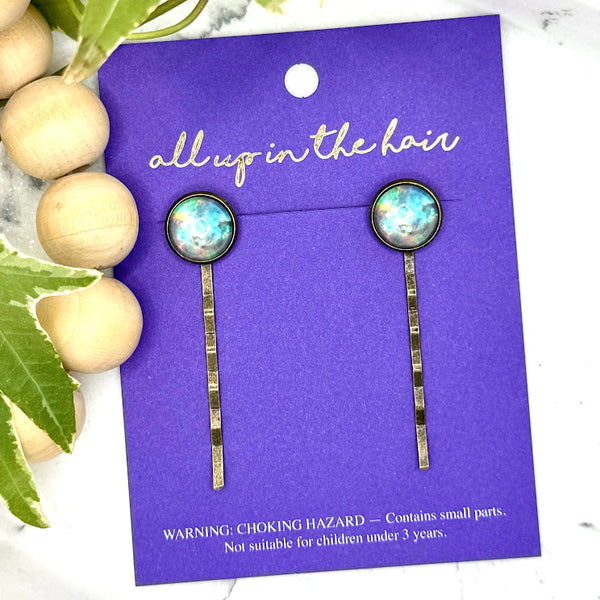 All Up In The Hair | Online Accessory Boutique Located in Mooresville, NC | Two Teal Galaxy Bobby Pins on an indigo colored, All Up In The Hair branded pacakging card. The card is laying on a white marble background alongside a wood bead garland and ivy leaves.
