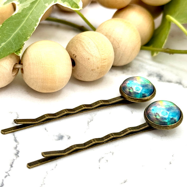 All Up In The Hair | Online Accessory Boutique Located in Mooresville, NC | Side view of two Teal Galaxy Bobby Pins laying on a white marble background alongside a wood bead garland and ivy leaves.