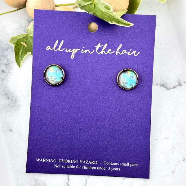 All Up In The Hair | Online Accessory Boutique Located in Mooresville, NC | Two Teal Galaxy Earrings on an indigo colored, All Up In The Hair branded packaging card. The card is laying on a white marble background.