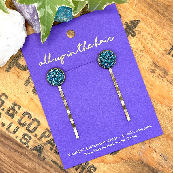 All Up In The Hair | Online Accessory Boutique Located in Mooresville, NC | Two Teal Glitter Druzy Bobby Pins on an indigo colored, All Up In The Hair branded packaging card. The card is laying on a wood background with black lettering. There are crystals and ivy leaves at the top of the image.