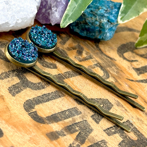 All Up In The Hair | Online Accessory Boutique Located in Mooresville, NC | Side view of two Teal Glitter Druzy Bobby Pins laying on a wood background with black lettering. There are crystals and ivy leaves at the top of the image.