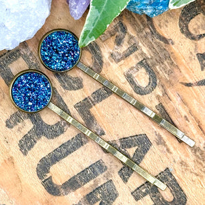 All Up In The Hair | Online Accessory Boutique Located in Mooresville, NC | Two glittery dark teal druzy bobby pins laying diagonally on a wood background with black lettering. There are crystals and ivy leaves at the top of the image.