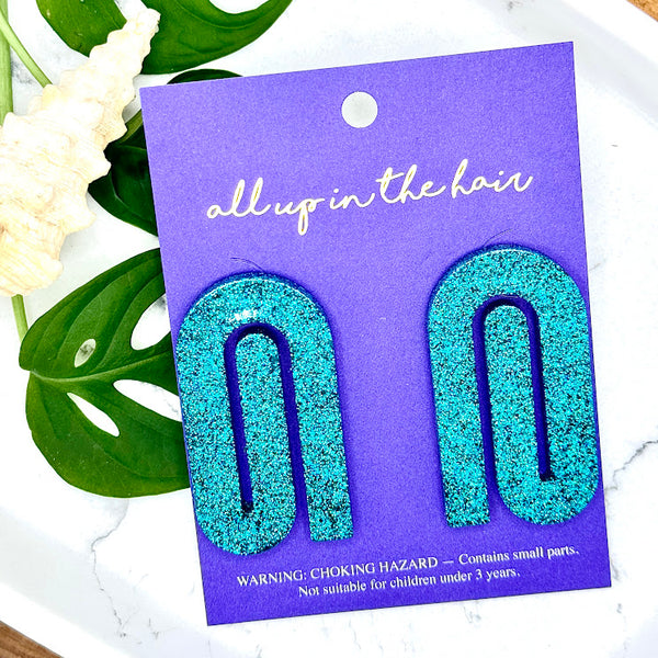 All Up In The Hair | Online Accessory Boutique Located in Mooresville, NC | Two teal paperclip earrings on an indigo backer card. The card is laying on two monstera leaves on a white background.