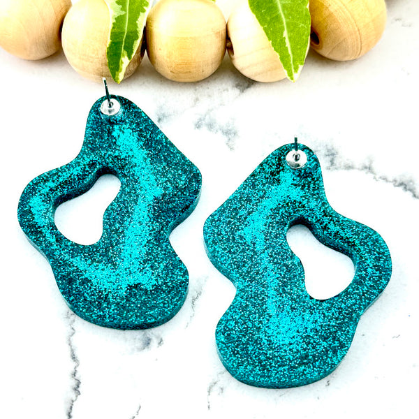 All Up In The Hair | Online Accessory Boutique Located in Mooresville, NC | Back of our Teal Wave Earrings laying on a white marble background. There is a wood bead garland and ivy leaves laying behind the earrings.