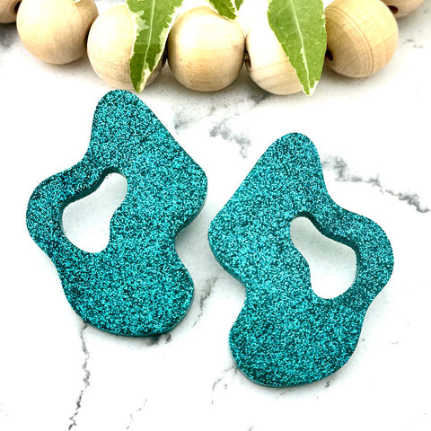 All Up In The Hair | Online Accessory Boutique Located in Mooresville, NC | Two Teal Wave Earrings on a white marble background. There is a wood bead garland and ivy leaves behind the earrings.