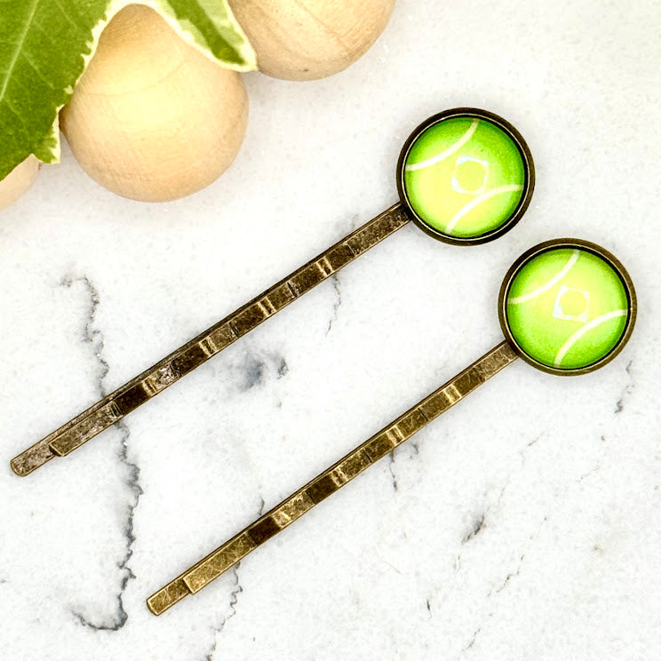 All Up In The Hair | Online Accessory Boutique Located in Mooresville, NC | Two Tennis Ball Bobby Pins laying diagonally on a wood background alongside a wood bead garland and ivy leaves. 