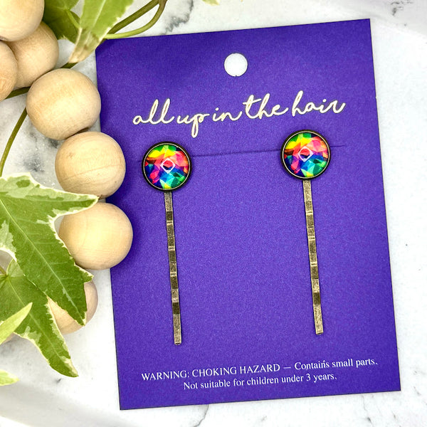 All Up In The Hair | Online Accessory Boutique Located in Mooresville, NC | Two Tie Dye Bobby Pins on an indigo colored, All Up In The Hair branded packaging card. The card is laying on a white marble background alongside a wood bead garland and ivy leaves.