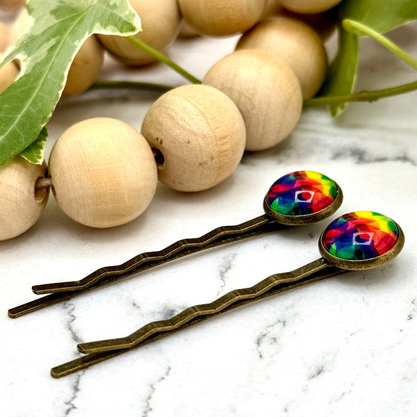 All Up In The Hair | Online Accessory Boutique Located in Mooresville, NC | Side view of two Tie Dye Bobby Pins on a white marble background alongside a wood bead garland and ivy leaves.