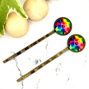 All Up In The Hair | Online Accessory Boutique Located in Mooresville, NC | Two Tie Dye Bobby Pins laying diagonally on a white marble background alongside a wood bead garland and ivy leaves.