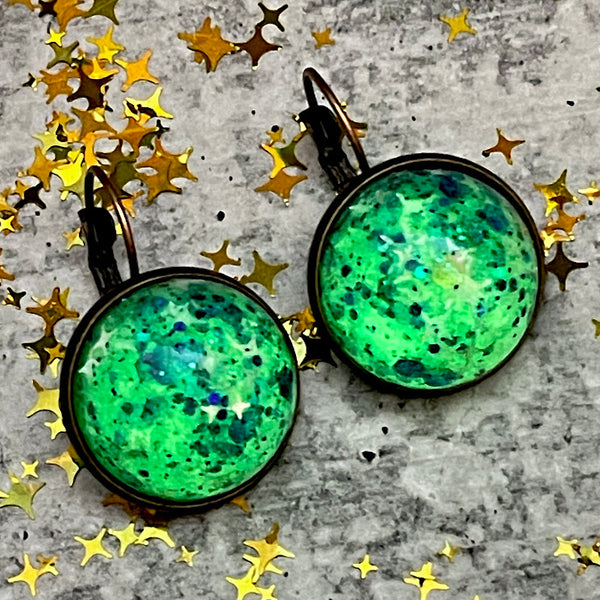 All Up In The Hair | Online Accessory Boutique Located in Mooresville, NC | Two quarter size dangle earrings glowing in lime green laying on a grey background, surrounded by gold star glitter.