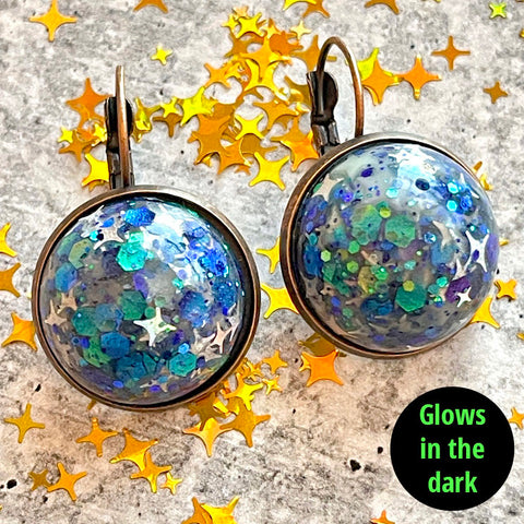 All Up In The Hair | Online Accessory Boutique Located in Mooresville, NC | Two quarter sized dangle earrings made with blue, teal and silver star glitter, laying on a grey background, surrounded by gold star glitter. In the bottom right hand corner is a black circle with the words "glow in the dark" in lime green writing.