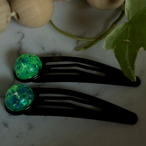 All Up In The Hair | Online Accessory Boutique Located in Mooresville, NC | Titan Snap Barrettes glowing green on a white marble background. At the top of the image is a wood bead garland and ivy leaves.