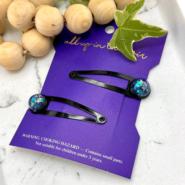 All Up In The Hair | Online Accessory Boutique Located in Mooresville, NC | Two Titan Snap Barrettes on an indigo colored, All Up In The Hair packaging card. The card is laying on a white marble background. At the top of the image is a wood bead garland and ivy leaves.