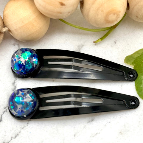 All Up In The Hair | Online Accessory Boutique Located in Mooresville, NC | Two Titan Snap Barrettes on a white marble background. The barrettes are black with a grey and blue cabochon. At the top of the image is a wood bead garland and ivy leaves.