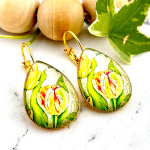 All Up In The Hair | Online Accessory Boutique Located in Mooresville, NC | Two teardrop earrings with a yellow tulip cabochon on a white marble background. Behind the earrings is a wood bead garland and ivy leaves.