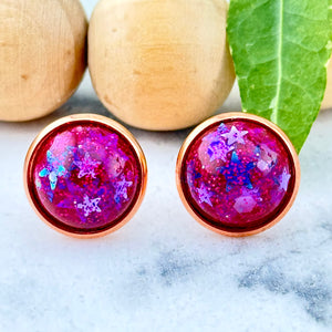 All Up In The Hair | Online Accessory Boutique Located in Mooresville, NC | Two pink, glittery studs containing tons of blue and silver stars on a white marble background. Behind the earrings is a wood bead garland and ivy leaves.
