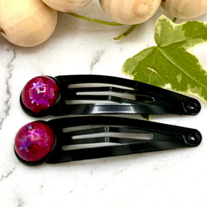 All Up In The Hair | Online Accessory Boutique Located in Mooresville, NC | Two black barrettes with bright pink cabochons on a white marble background. At the top of the image is a wood bead garland and ivy leaves.