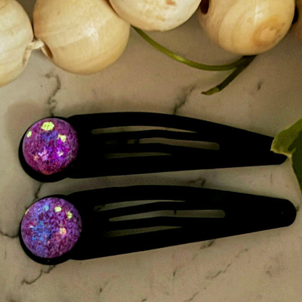 All Up In The Hair | Online Accessory Boutique Located in Mooresville, NC | Two black snap barrettes with glowing periwinkle cabochons are laying on a grey background, surrounded by gold star glitter.