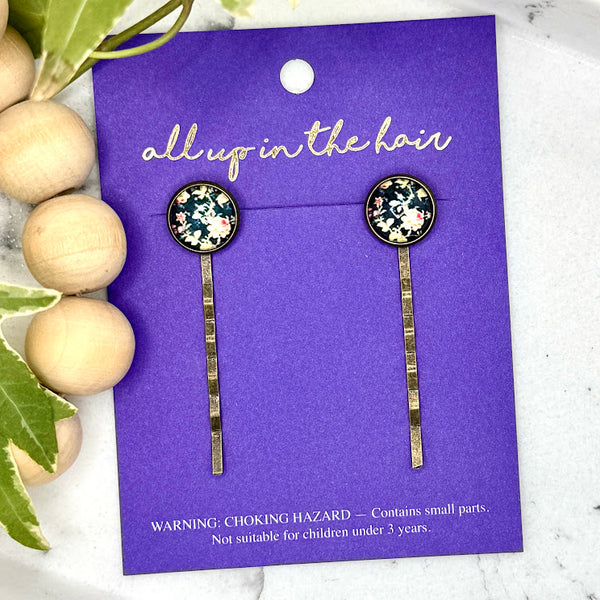 All Up In The Hair | Online Accessory Boutique Located in Mooresville, NC | Two Vintage Floral Bobby Pins on an indigo colored, All Up In The Hair branded packaging card. The card is laying on a white marble background alongside a wood bead garland and ivy leaves.