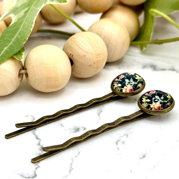 All Up In The Hair | Online Accessory Boutique Located in Mooresville, NC | Side view of two Vintage Floral Bobby Pins laying on a white marble background alongside a wood bead garland and ivy leaves.