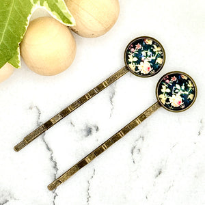 All Up In The Hair | Online Accessory Boutique Located in Mooresville, NC | Two Vintage Floral Bobby Pins laying diagonally on a white marble background alongside a wood bead garland and ivy leaves.