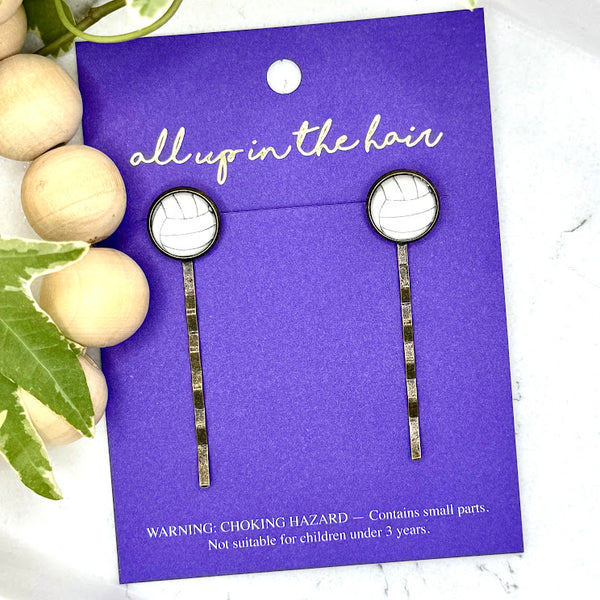 All Up In The Hair | Online Accessory Boutique Located in Mooresville, NC | Two Volleyball Bobby Pins on an indigo colored, All Up In The Hair branded packaging card. The card is laying on a white marble background alongside a wood bead garland and ivy leaves.