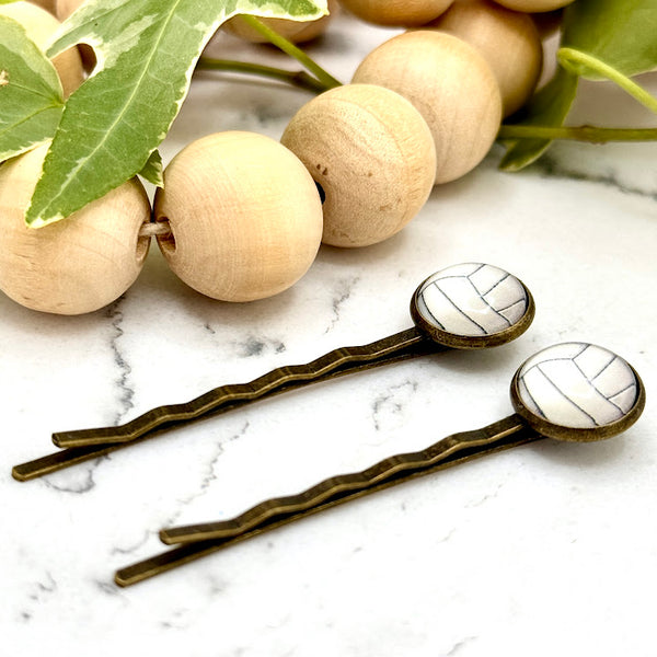 All Up In The Hair | Online Accessory Boutique Located in Mooresville, NC | Side view of two Volleyball Bobby Pins on a white marble background alongside a wood bead garland and ivy leaves.