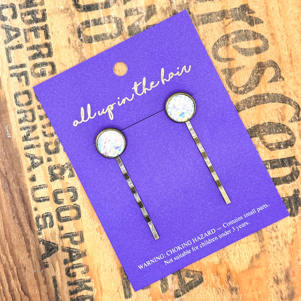 All Up In The Hair | Online Accessory Boutique Located in Mooresville, NC | Two White Rainbow Mermaid Bobby Pins on an indigo colored, All Up In The Hair branded packaging card. The card is laying on a wood background with black lettering.